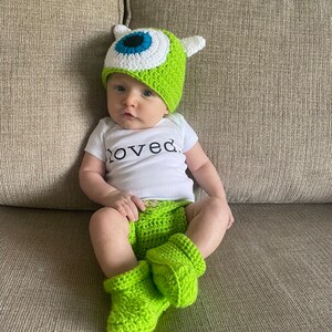 May include: A baby wearing a green crocheted hat with a large blue and white eye design, matching green diaper cover, and booties. The baby is also wearing a white onesie with the word "loved" printed on it.
