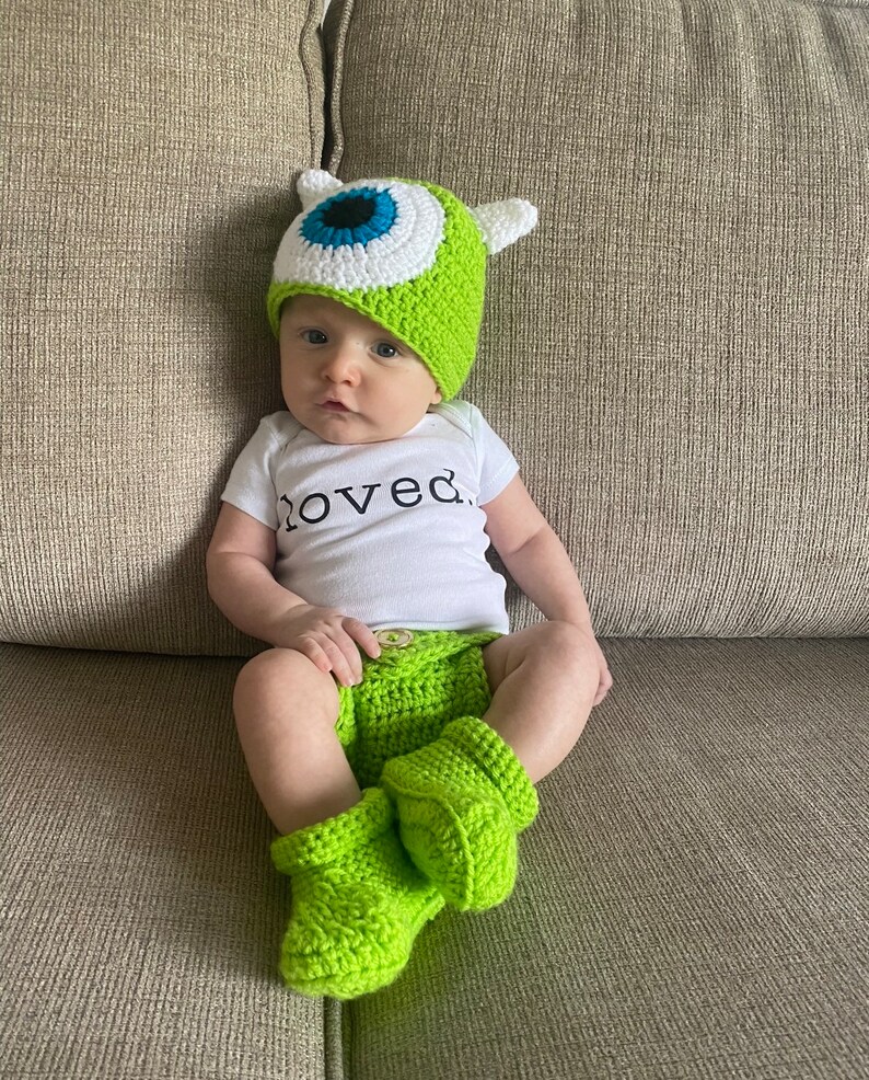 May include: A baby wearing a green crocheted hat with a large blue and white eye design, matching green diaper cover, and booties. The baby is also wearing a white onesie with the word "loved" printed on it.
