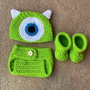 May include: A green crocheted baby outfit set, including a hat with white ears and a large eye design, a diaper cover, and a pair of booties. The set is made of a soft, textured yarn.