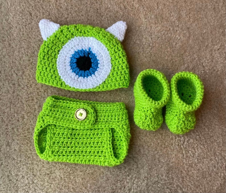 May include: A green crocheted baby outfit set, including a hat with white ears and a large eye design, a diaper cover, and a pair of booties. The set is made of a soft, textured yarn.