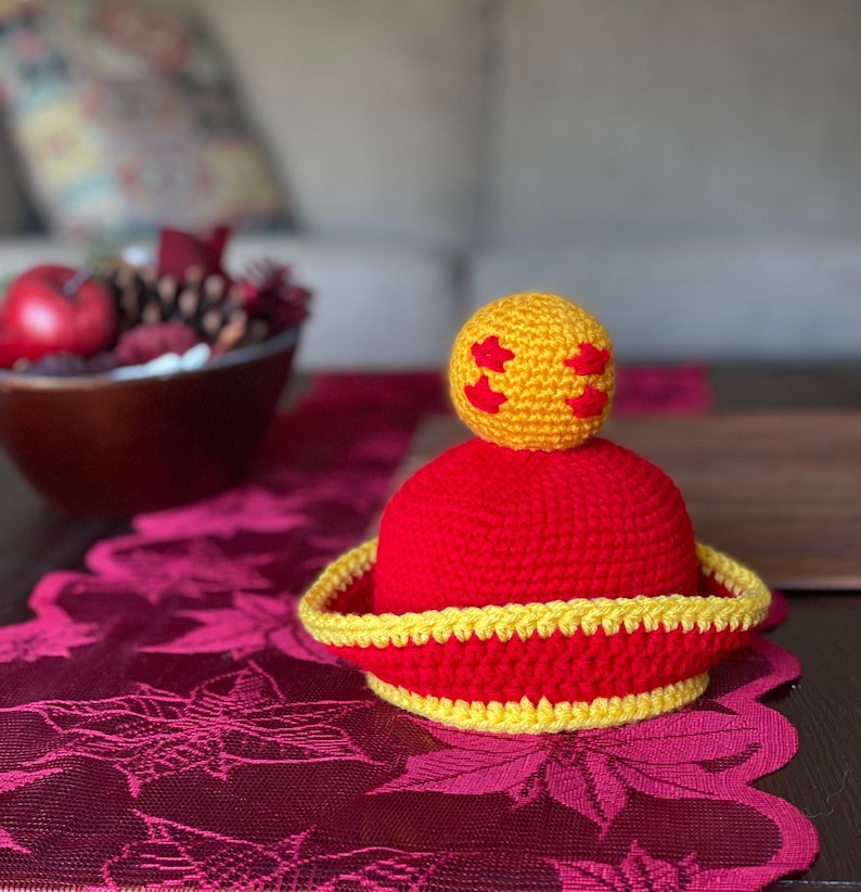 May include: A red and yellow crocheted hat with a yellow crocheted ball on top. The hat has a wide brim and the ball has a star pattern.