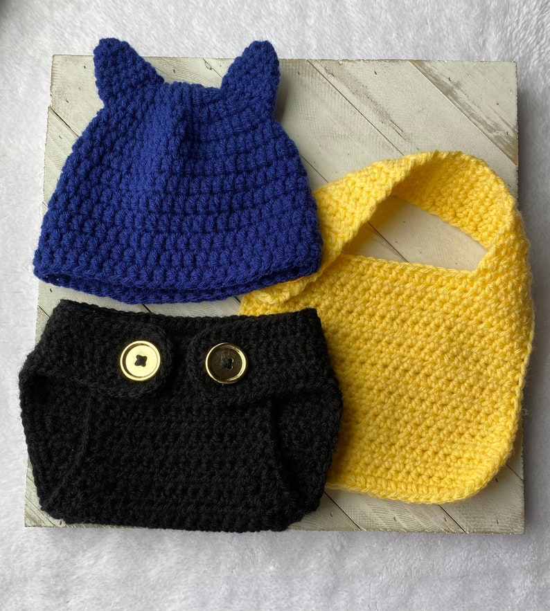May include: A crocheted baby outfit set in blue, black, and yellow. The set includes a hat with ears, a diaper cover, and a bib.