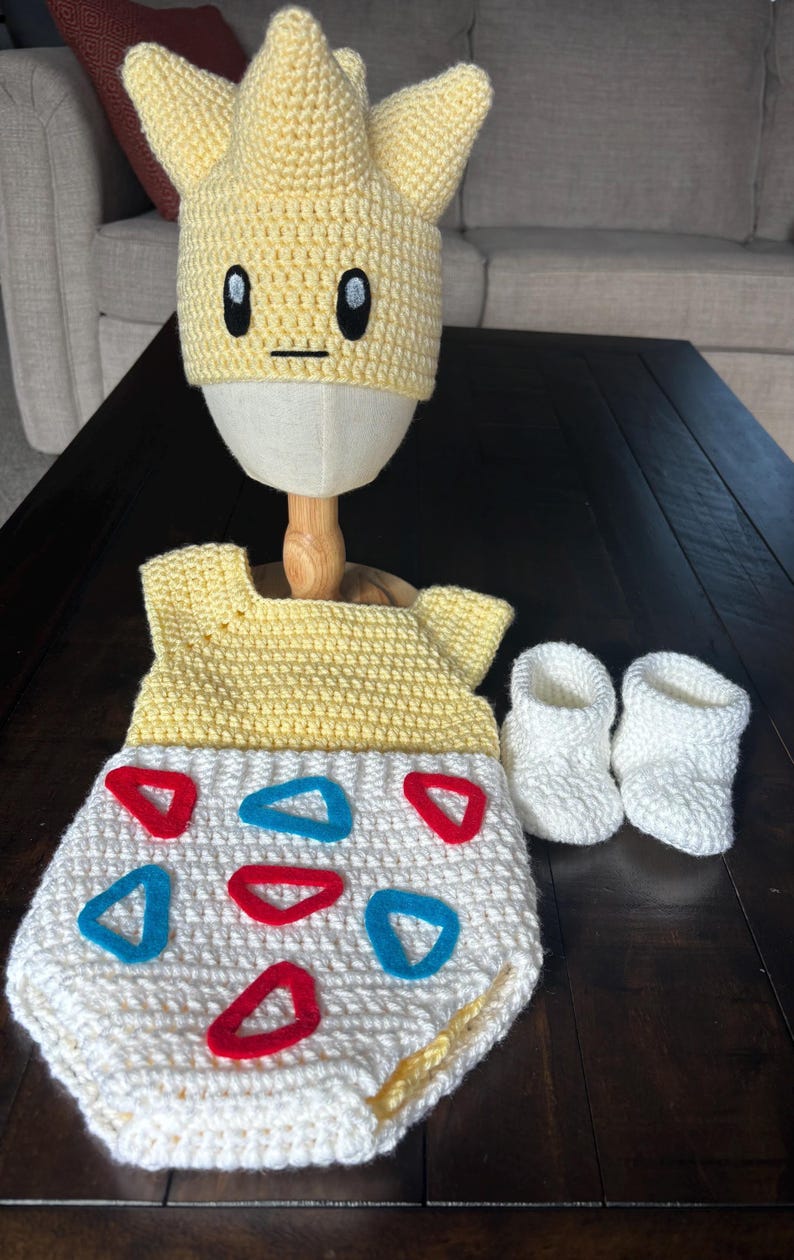K&ouml;nnte beinhalten: Handgefertigtes Baby-Set aus H&auml;kelarbeit in Gelb und Wei&szlig;, bestehend aus Hut, Strampler und Babyschuhen. Der Strampler hat rote und blaue Dreiecksakzente. Der Hut ist mit einem Cartoon-Gesicht gestaltet.