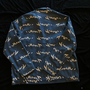 Op de afbeelding: Blauwe spijkerjas met een wit "Monch" scriptpatroon op de rug. De jas heeft een knoopsluiting en lange mouwen. De stof is een mediumgewicht denim.