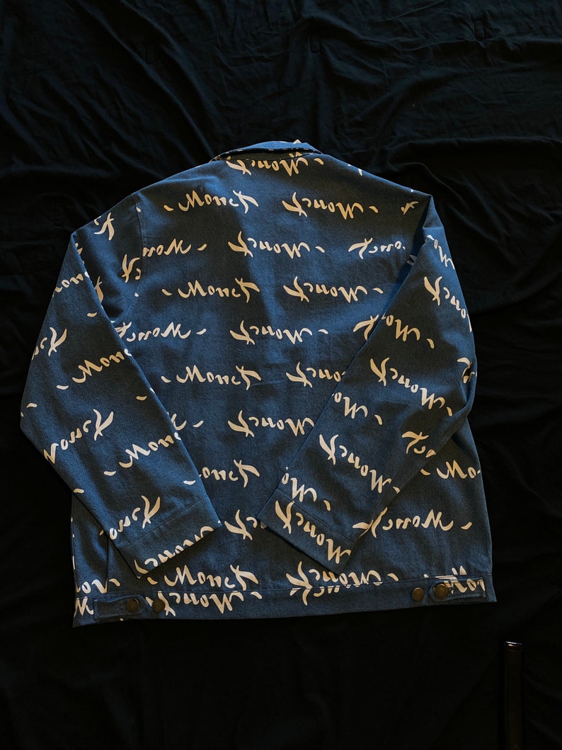 Op de afbeelding: Blauwe spijkerjas met een wit "Monch" scriptpatroon op de rug. De jas heeft een knoopsluiting en lange mouwen. De stof is een mediumgewicht denim.