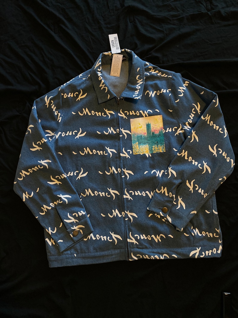 Op de afbeelding: Blauw spijkerjasje met het woord "Monet" in witte letters herhaald over de stof. Een kleine zak heeft een schilderij van een gebouw boven water. De jas heeft een ritssluiting en manchetten met knopen.