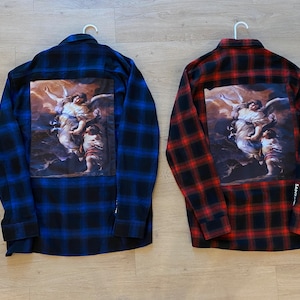 May include: Two long-sleeved flannel shirts with a black and white checked pattern. One shirt is blue and the other is red. Both shirts feature a large image of an angel with wings on the back. The image is printed on a white background.