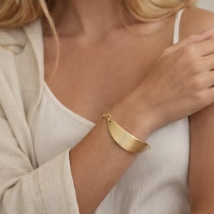 May include: A gold-colored bracelet with a curved, elongated design. The bracelet is made of metal and has a smooth, matte finish. It is connected by a small chain on each end.