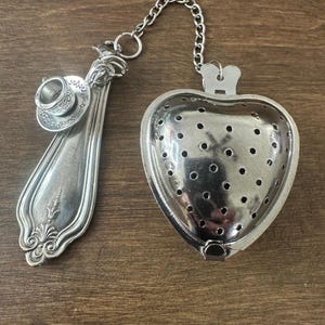 May include: A silver tea infuser with a heart-shaped basket and a decorative spoon handle. The spoon handle features a miniature teacup and saucer. A chain connects the basket to the spoon.