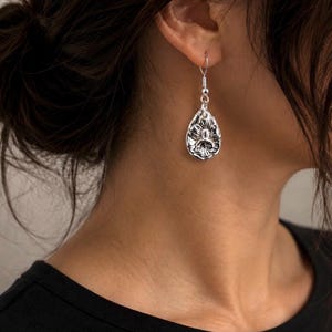 May include: A silver-toned drop earring featuring a teardrop shape with an embossed floral design. The earring hangs from a simple silver-toned hook wire, showcasing a polished metallic finish and intricate petal details on the surface.