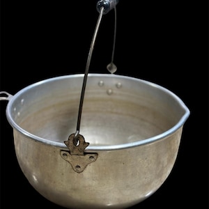 May include: A vintage aluminum cooking pot with a black handle and a metal hanging loop. The pot has a rounded shape with a pouring spout and is set against a black background. The pot is likely used for cooking or boiling.