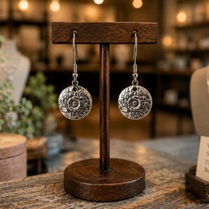 May include: A pair of silver-toned dangle earrings featuring a circular charm with an embossed floral pattern. The earrings hang from simple metal hooks and are displayed on a dark wooden T-shaped stand. A small card nearby reads Vintage Stories Timeless Beauty.