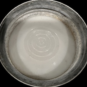 May include: Close-up of a white, concave object with concentric circles in the center. The object is surrounded by a silver-colored, ridged rim. The rim has a slightly weathered appearance. The object appears to be a lid or cap.