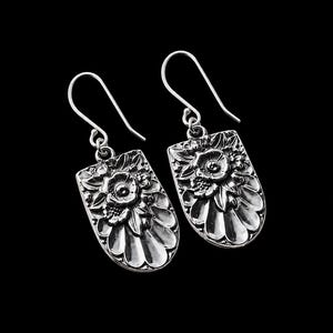 May include: A pair of silver-toned drop earrings featuring a rectangular shape with rounded bottom edges. The surface displays a raised, detailed floral relief pattern with deep oxidized accents, suspended from simple curved silver-toned ear wires.