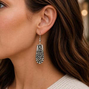 May include: A silver-toned dangle earring featuring an elongated, spoon-shaped pendant with an intricate, embossed floral pattern. The metal surface displays a dark, oxidized finish within the recessed areas to highlight the detailed botanical design.