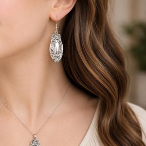May include: A silver-toned jewelry set consisting of a pendant necklace and matching drop earrings. Each piece features an elongated oval shape with intricate floral relief detailing at the top and bottom, surrounding a smooth, polished center.