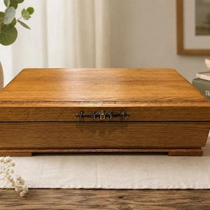 May include: A rectangular wooden box with a natural oak grain finish and a smooth, flat lid. The box features a dark metal decorative latch centered on the front panel and rests on a slightly elevated base with small feet at each corner.