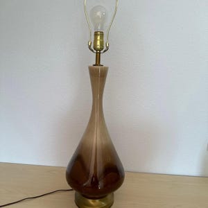 May include: A vintage table lamp with a teardrop-shaped ceramic base in shades of brown and beige. The lamp has a brass base and a brass lamp holder, with a light bulb installed. The lamp is approximately 20 inches tall.