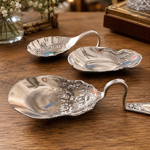 May include: Three silver-toned metal spoons with wide, shallow, shell-shaped bowls and ornate floral embossed handles. The spoons feature curved, elevated stems and detailed rose patterns on the bowls and handles, reflecting a metallic sheen.