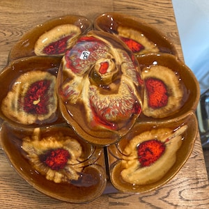May include: A vintage ceramic serving dish with a central swirl design and six surrounding sections. The dish features a glossy finish with brown, tan, and red hues. The design resembles a seashell or flower.