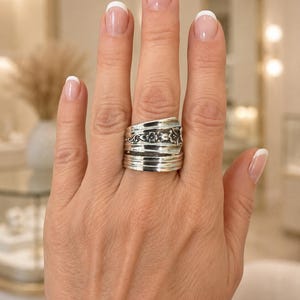 May include: A silver spoon ring featuring an intricate floral pattern on the upper band and three horizontal grooves on the lower band. The metal has a polished finish with darkened recessed areas that highlight the embossed botanical details.