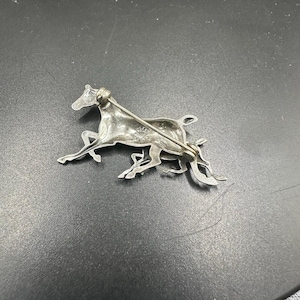 May include: Silver tone horse brooch with a detailed design. The brooch is in the shape of a horse running with its tail flowing behind it.