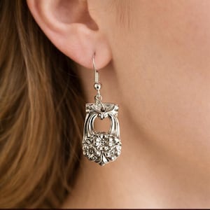 May include: Silver-toned floral earrings. The earrings feature a detailed floral design at the bottom and a decorative top. The earrings are hanging from a silver fish hook style ear wire. The earrings are worn on a person's ear.