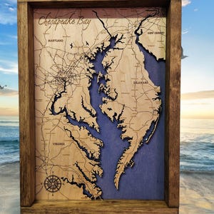 May include: Wooden framed map of Chesapeake Bay. The map is laser cut from light-colored wood, with a blue background representing the water. The map includes the names of states and cities. The words "Chesapeake Bay" are written in cursive at the top.