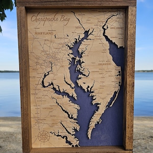 May include: Wooden framed map of the Chesapeake Bay. The map is laser-cut from light-colored wood, showing the bay's outline and surrounding states. The background is a dark blue, and the frame is a dark brown. The words "Chesapeake Bay" are at the top.