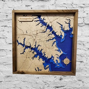 May include: A wooden framed map of Annapolis, Maryland, with a layered design showing the city's layout and waterways in shades of blue and beige. A compass rose is in the lower right corner, and the word "Annapolis" is at the top.