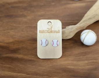May include: Baseball stud earrings on a wooden display card. The earrings are white with red stitching, resembling baseballs. The card is branded with the text "RUSTIC HALO". The background is a wooden surface.