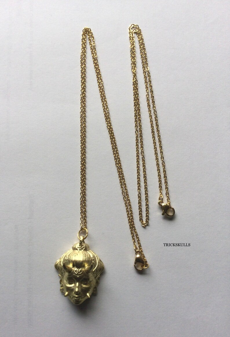 May include: A gold chain necklace with a pendant featuring a detailed, stylized face with horns. The pendant is a 3D sculpture with a textured surface. The chain is a delicate, linked chain.