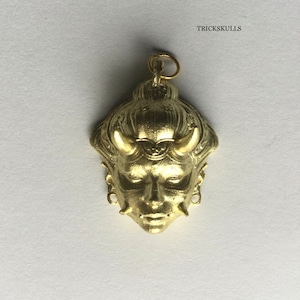 May include: A gold-colored pendant featuring a detailed, stylized face with horns and intricate designs. The pendant has a loop at the top for attaching to a chain.