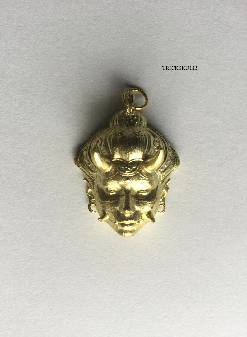 May include: A gold-colored pendant featuring a detailed, stylized face with horns and intricate designs. The pendant has a loop at the top for attaching to a chain.