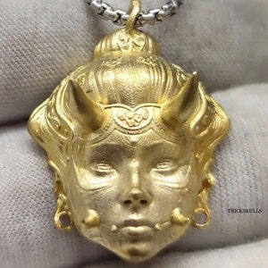 May include: Gold pendant necklace featuring a detailed, stylized face with horns and intricate designs. The pendant is attached to a silver chain.