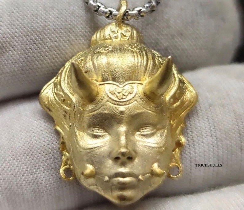 May include: Gold pendant necklace featuring a detailed, stylized face with horns and intricate designs. The pendant is attached to a silver chain.