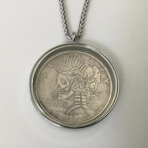 May include: Silver pendant necklace featuring a skull with a crown of thorns, surrounded by a silver frame. The pendant reads "LIBERTY", "IN GOD WE TRUST", and "1913".