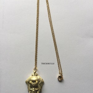 May include: A gold chain necklace with a pendant featuring a detailed, stylized face with horns. The pendant is a 3D sculpture with intricate details. The text "TRICKSKULLS" is visible in the image.
