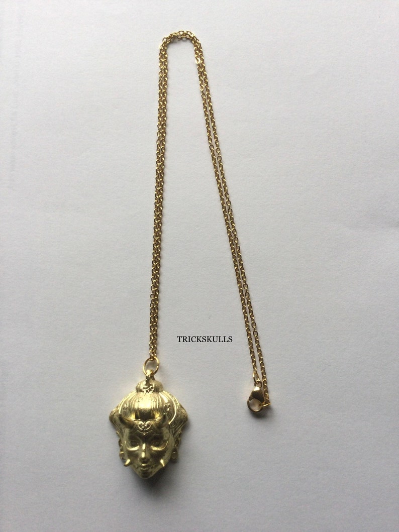 May include: A gold chain necklace with a pendant featuring a detailed, stylized face with horns. The pendant is a 3D sculpture with intricate details. The text "TRICKSKULLS" is visible in the image.