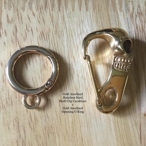 May include: Two gold-colored metal accessories are displayed on a wooden surface. One is a circular ring with a clasp, and the other is a skull-shaped clip carabiner. The text in the image reads: Gold Anodized Stainless Steel Skull Clip Carabiner + Gold Anodized Opening O Ring.