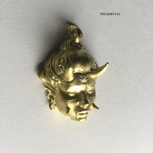 May include: Gold-colored pendant featuring a detailed, three-dimensional sculpture of a woman's face with horns. The pendant has a loop at the top for attaching to a chain.