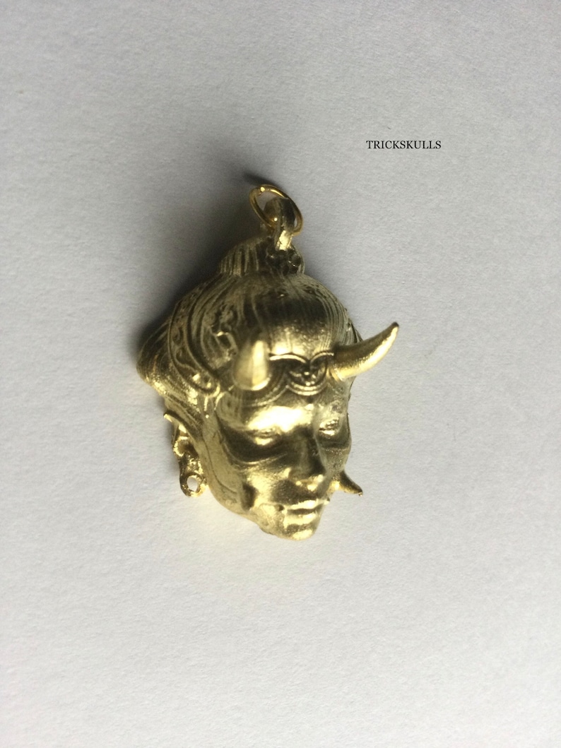 May include: Gold-colored pendant featuring a detailed, three-dimensional sculpture of a woman's face with horns. The pendant has a loop at the top for attaching to a chain.