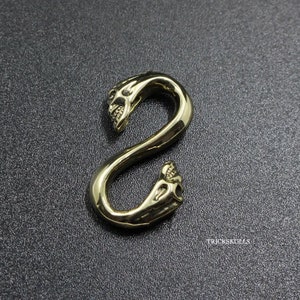 May include: A gold-colored metal skull-shaped S-hook. The hook is designed with two skull heads, one at each end of the S-shape.