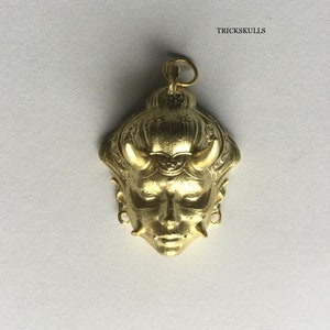 May include: A gold-colored pendant featuring a detailed, stylized face with horns and intricate details. The pendant is a traditional Japanese demon mask.