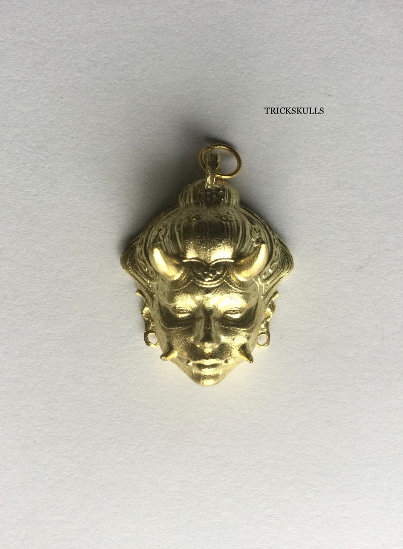 May include: A gold-colored pendant featuring a detailed, stylized face with horns and intricate details. The pendant is a traditional Japanese demon mask.