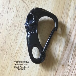 May include: Black anodized stainless steel skull clip. The clip features a skull design on one end and a spring-loaded gate on the other. The text "TRICKSKULLS Stainless Steel Black Anodized Skull Clip" is visible.