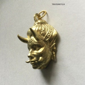 May include: Gold-colored pendant featuring a detailed, stylized face with horns and intricate designs. The pendant has a loop at the top for attaching to a chain.