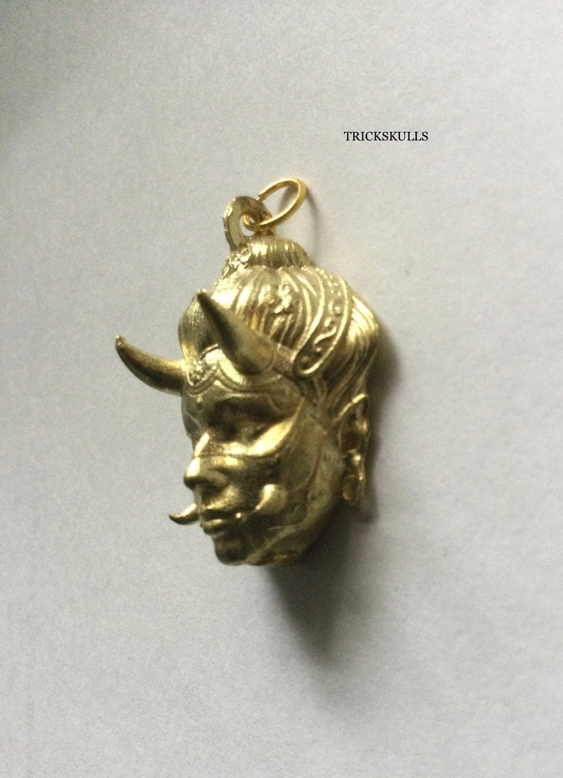 May include: Gold-colored pendant featuring a detailed, stylized face with horns and intricate designs. The pendant has a loop at the top for attaching to a chain.