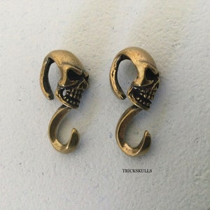 May include: Two gold-toned skull-shaped hooks. The hooks are detailed with a skull face and have a curved shape.