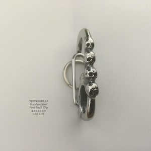 May include: A stainless steel clip with a series of four skull designs. The clip is 1.61 inches wide and 0.71 inches tall. The text "TRICKSKULLS" is visible, along with the product name and dimensions.